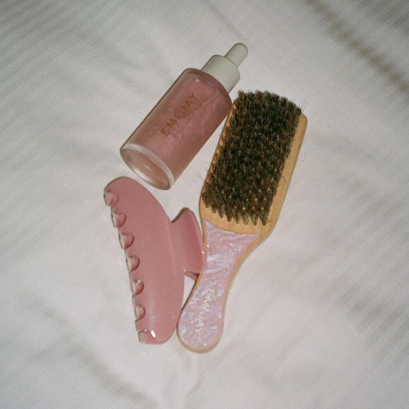 Pink glass facial roller, gua sha stone, boar bristle brush and EMMLAY serum bottle on white fabric background