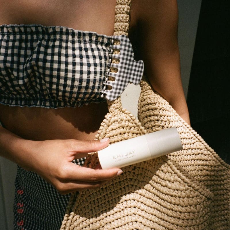 Woman holding cream-colored woven straw handbag with beaded strap and white product tube, wearing gingham top