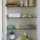 Glass bathroom shelves displaying spa essentials including towels, hair clips, brushes, and skincare products in neutral tones