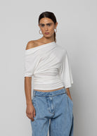 White off-shoulder draped top with ruched waist detail and short sleeves, paired with light blue wide-leg jeans.