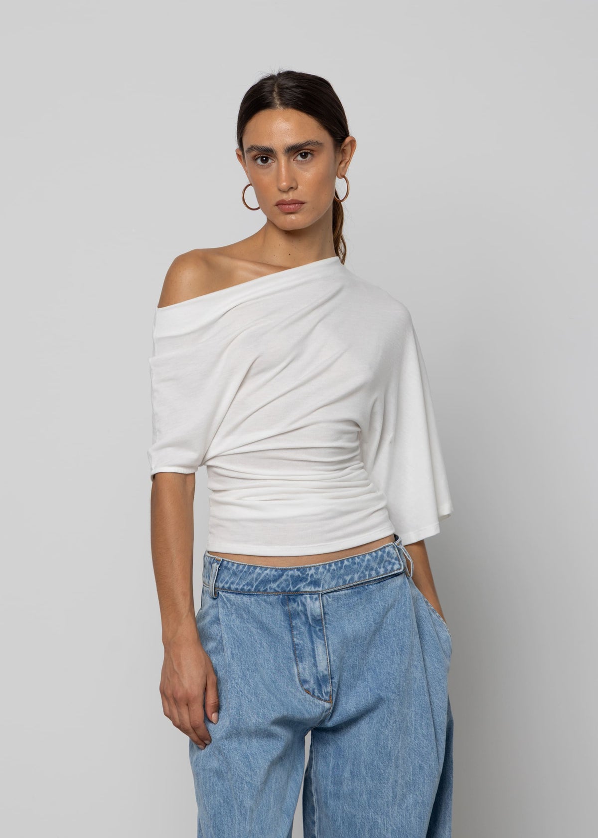 White off-shoulder draped top with ruched waist detail and short sleeves, paired with light blue wide-leg jeans.
