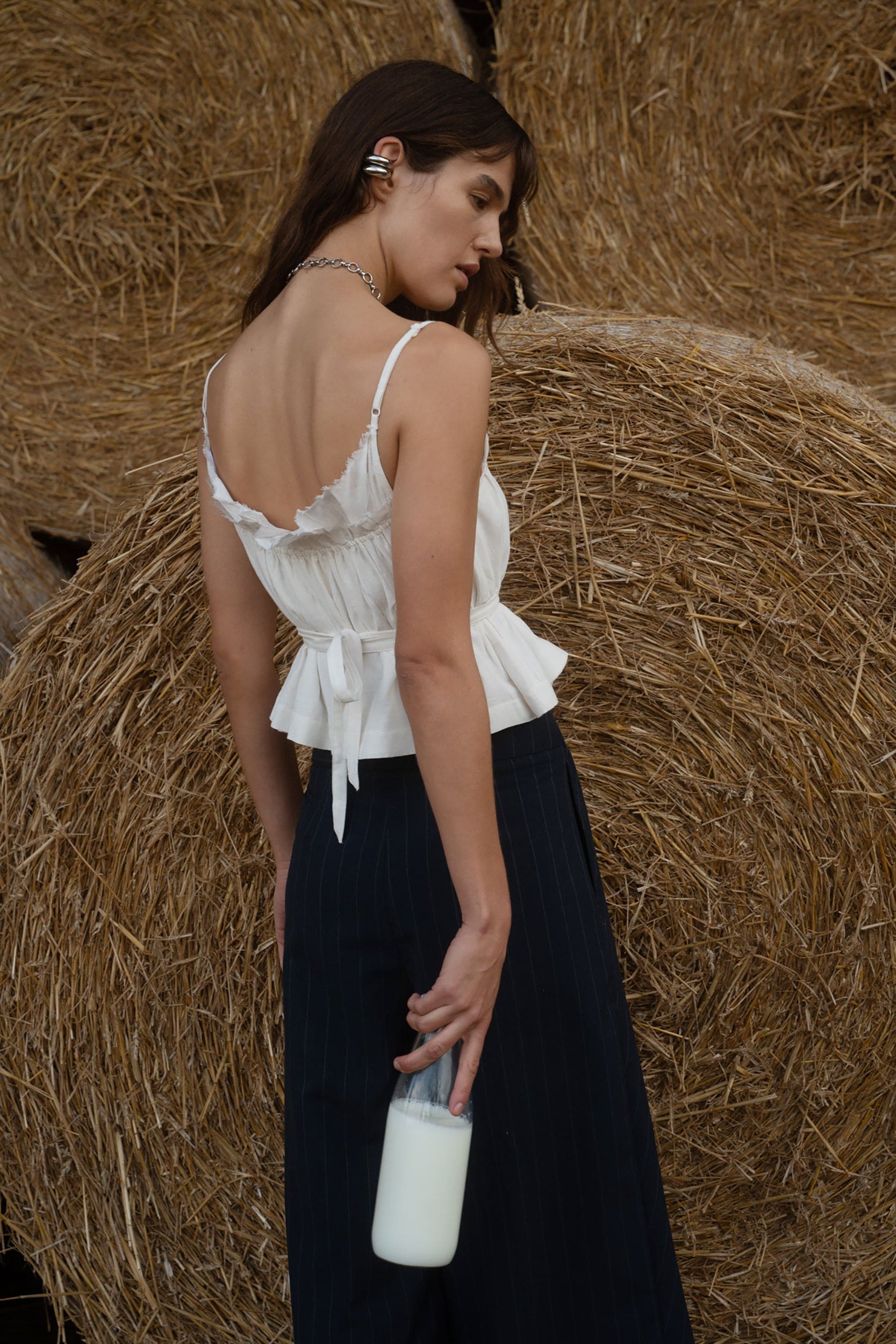 Woman in a white top and black skirt holding a bottle, standing in front of hay bales.