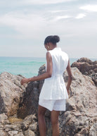 Woman in white sleeveless belted shirt dress standing on rocky coastline overlooking turquoise ocean water