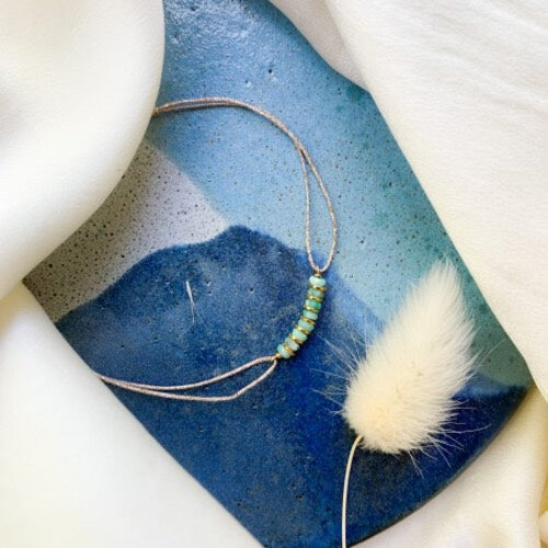 Delicate gold bracelet with green beads on watercolor blue ceramic dish with white fur accent
