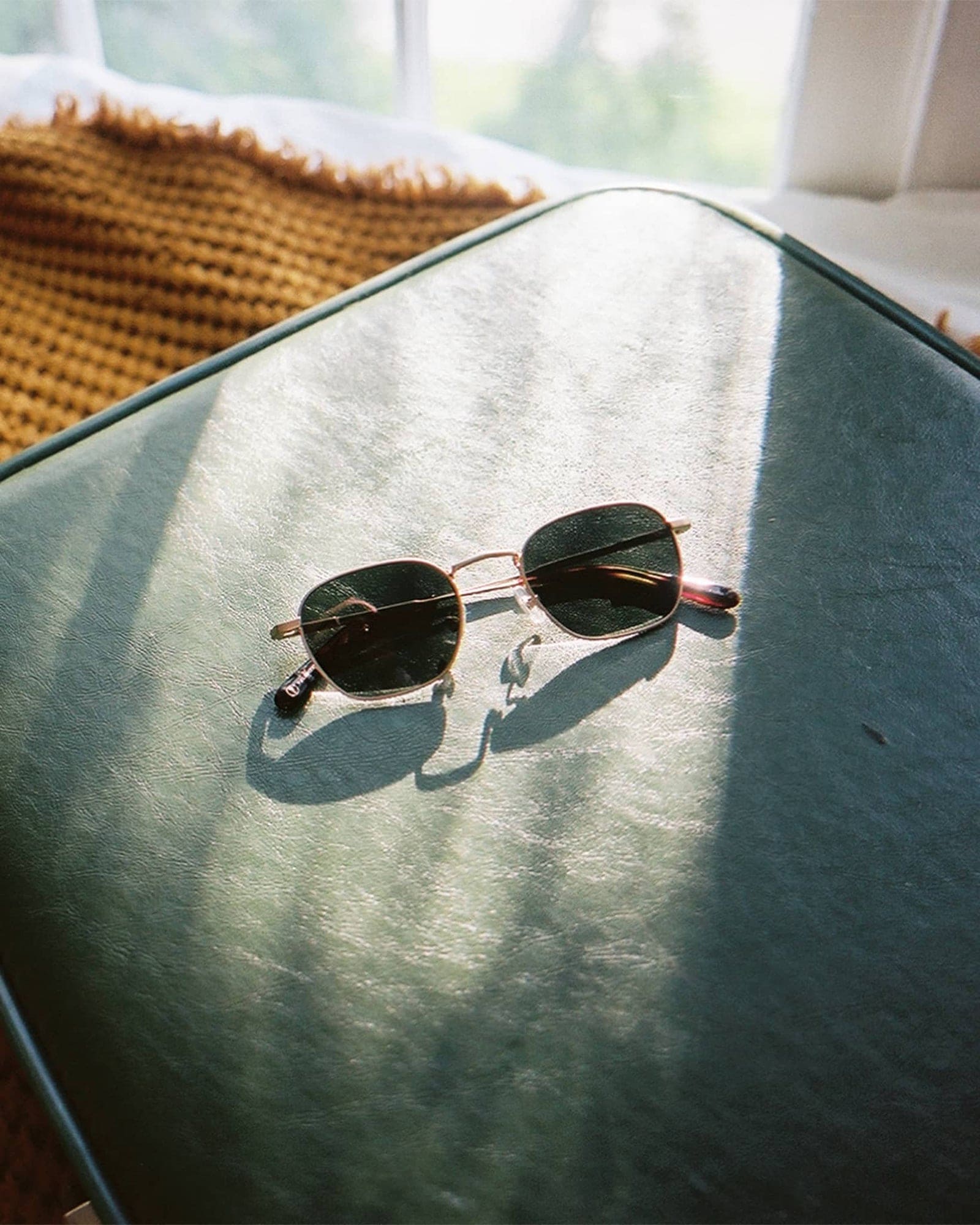 Round metal frame sunglasses with dark lenses and tortoiseshell temple tips resting on a sage green velvet cushion
