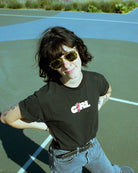 Person wearing black t-shirt with "GIRL" graphic print and skateboarder logo, styled with yellow sunglasses at skate park