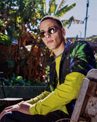 Young man wearing round sunglasses and colorful jacket with green palm leaf print over yellow shirt in tropical outdoor setting