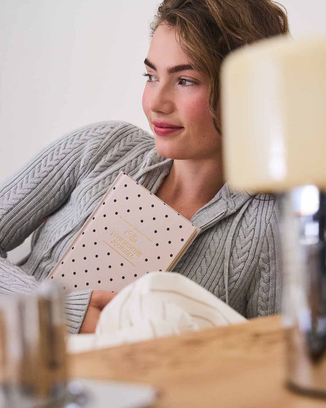 Person in gray cable knit sweater holding pink polka dot gratitude journal while relaxing with coffee at home