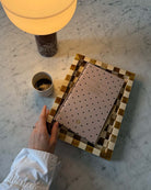 Checkered bone inlay tray in gold and cream tones holding a polka dot journal, displayed on marble surface with coffee cup