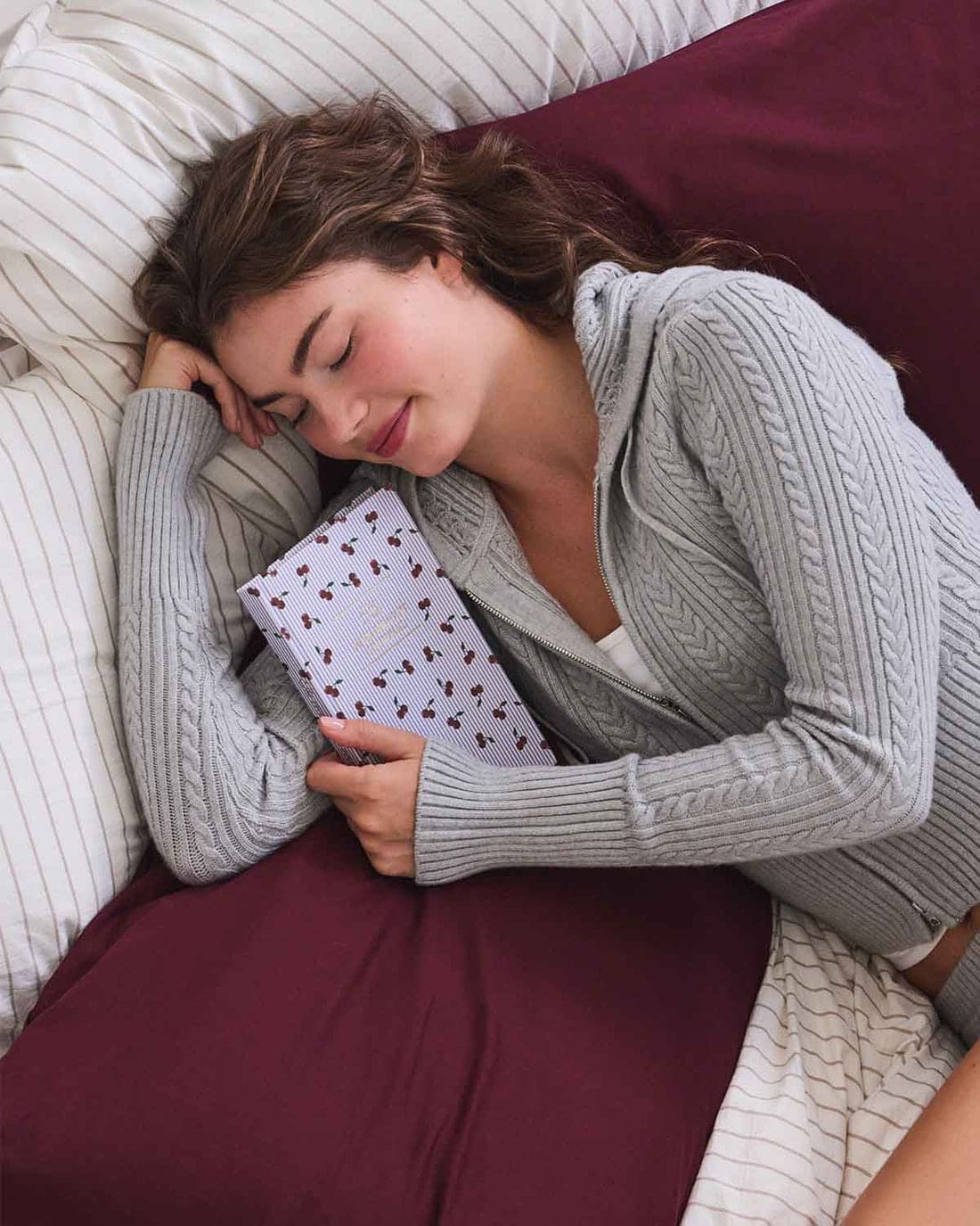 Lavender heating pad with cherry pattern held by person in gray sweater lying in bed with burgundy blanket