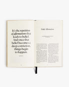 Open journal showing daily affirmation pages with black text on cream paper, featuring Muhammad Ali quote about repetition and belief