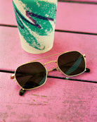 Gold-framed sunglasses with dark lenses on a pink wooden surface next to a colorful cup.