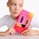 Person with bald head holding colorful gradient face mask package featuring pink and orange design with abstract shapes