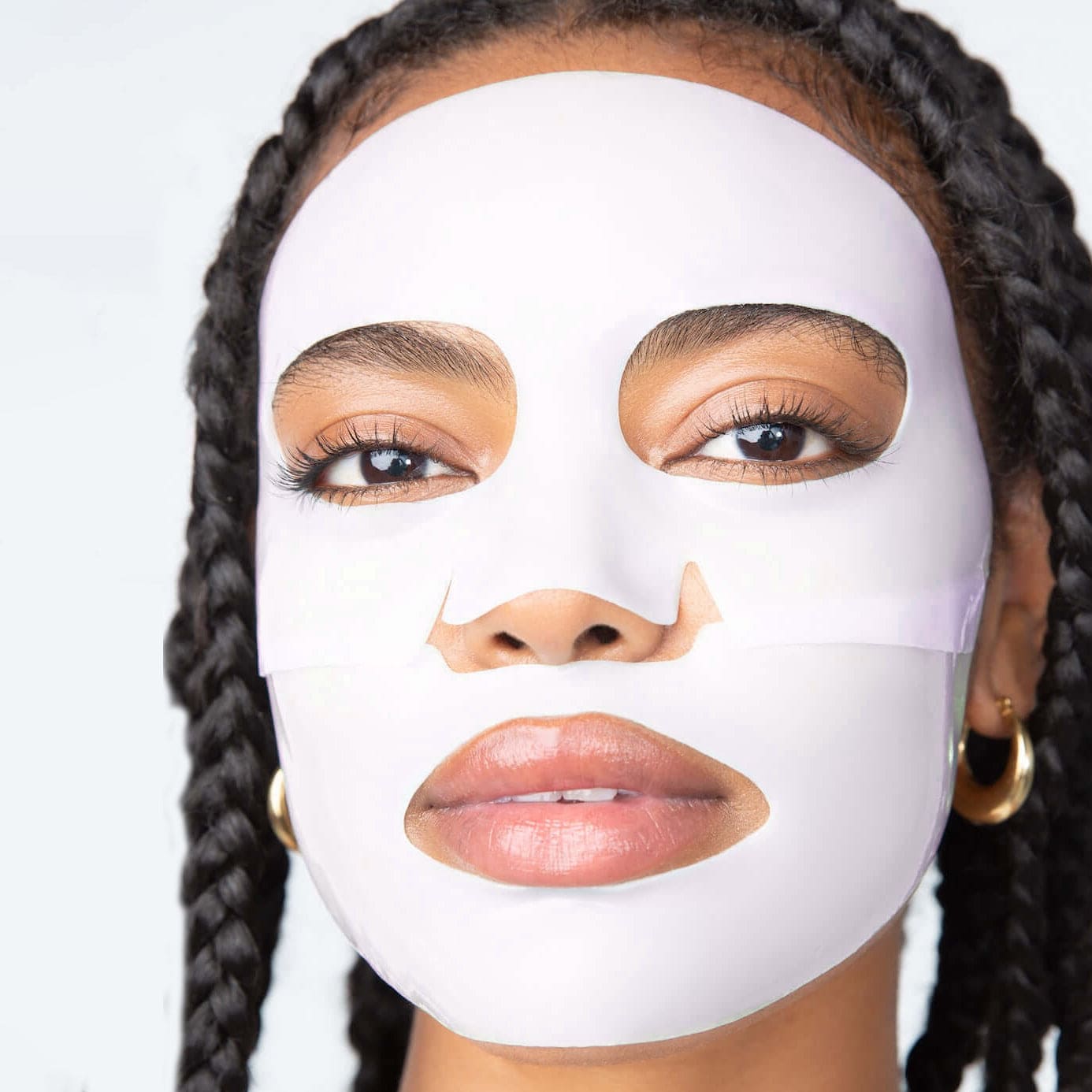 White sheet face mask on person with braided hair, showing cutouts for eyes, nose, and mouth for skincare treatment