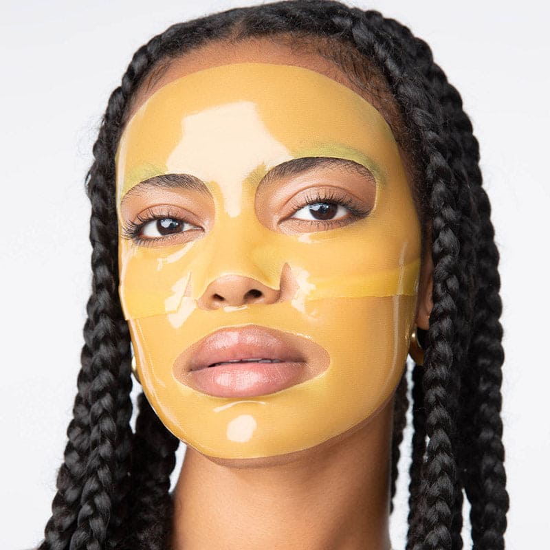 Woman wearing golden yellow hydrating sheet face mask with cutouts for eyes, nose, and mouth, long braided hairstyle