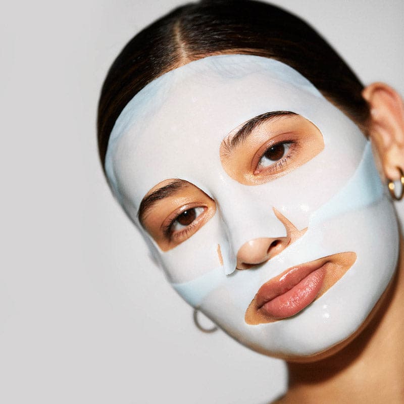 Woman wearing white sheet face mask with cutouts for eyes, nose, and mouth against neutral background