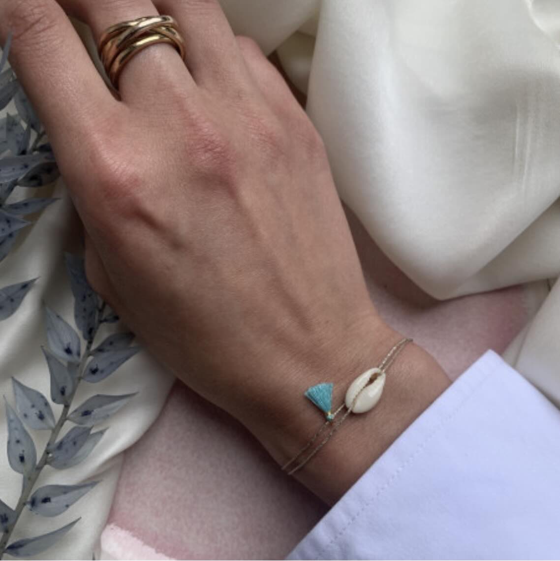 Delicate gold chain bracelet with turquoise tassel charm and cowrie shell on wrist, shown with matching stacked rings