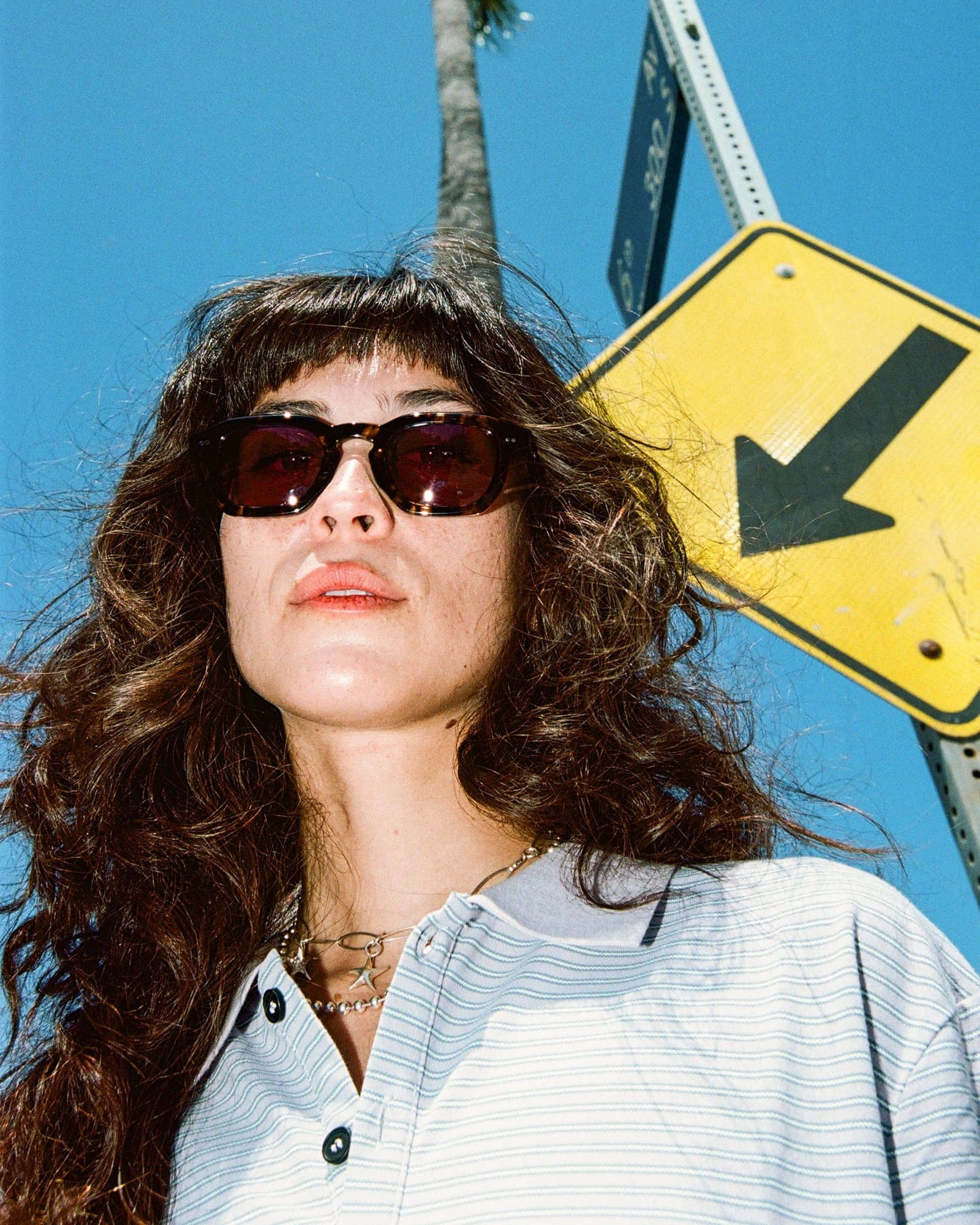 Woman with long wavy hair wearing dark tortoiseshell sunglasses and white striped shirt against blue sky with yellow road sign