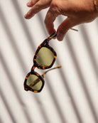 Tortoiseshell sunglasses with yellow-green tinted lenses held by hand against white fabric background