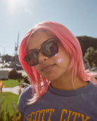 Person with pink hair wearing dark sunglasses and navy t-shirt outdoors on sunny day with green grass and trees in background
