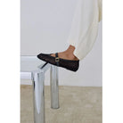 Black mesh flat shoes with gold buckle detail worn with white pants on chrome stool