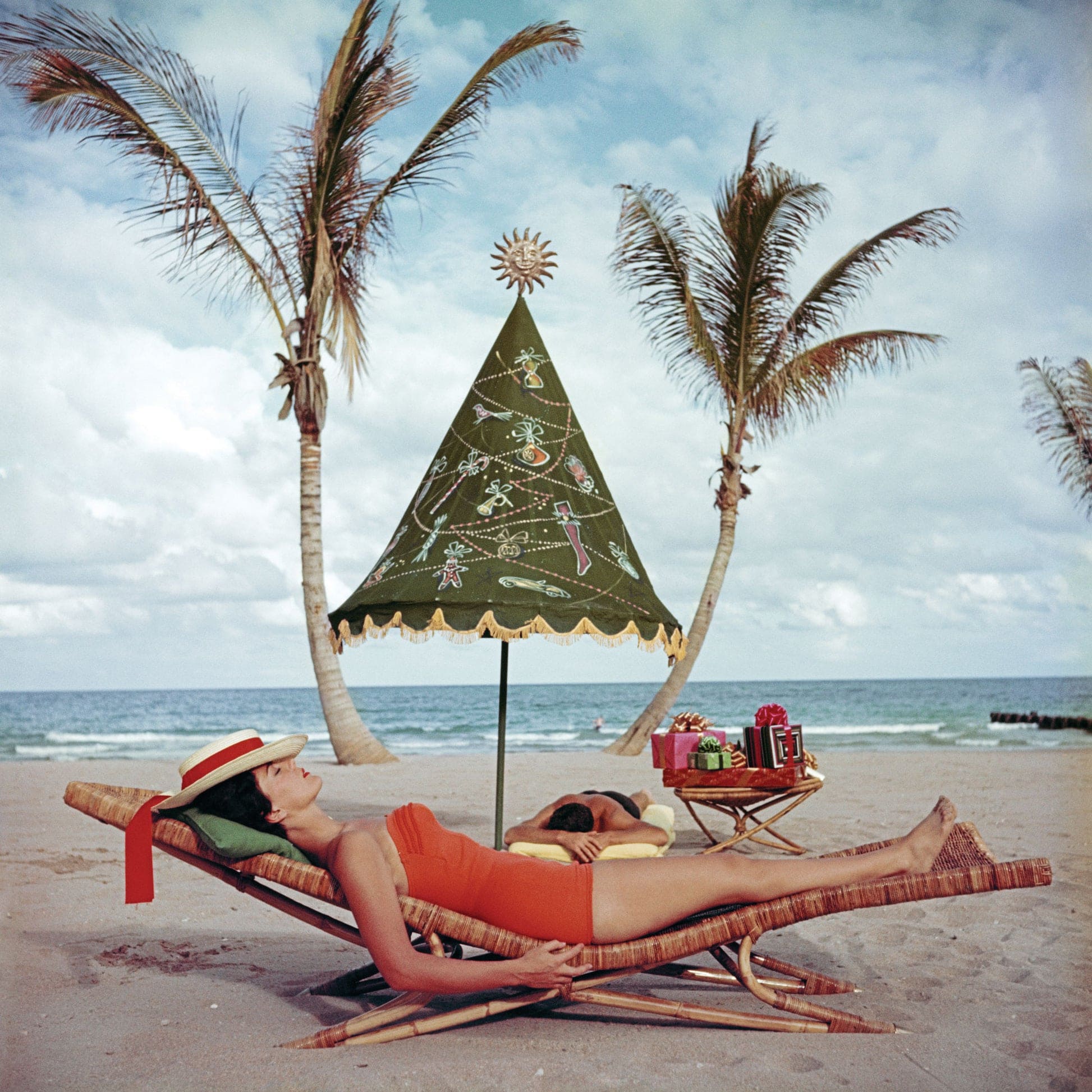 Woman relaxing on beach lounge chair under decorative green Christmas tree umbrella with palm trees and ocean backdrop