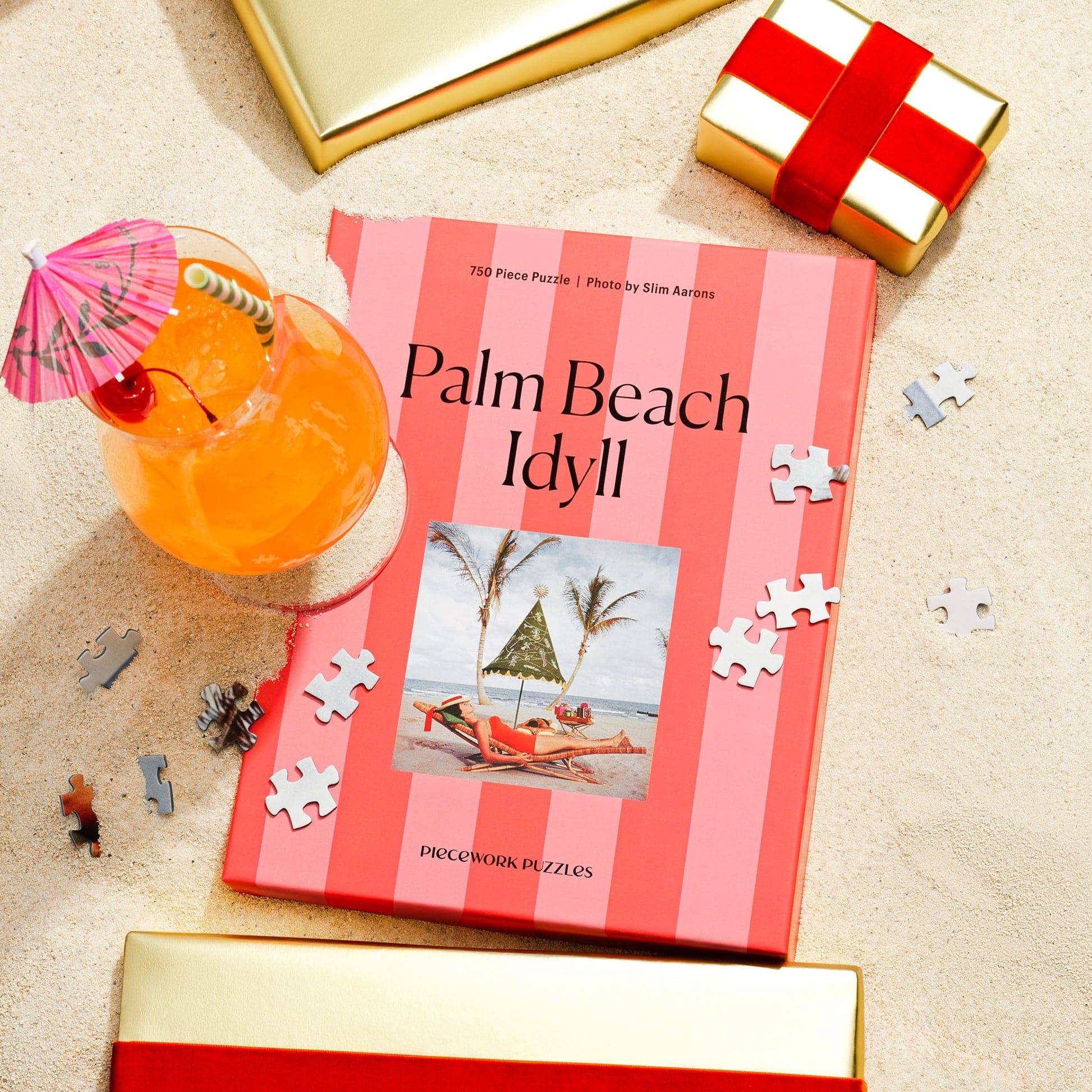 Palm Beach Idyll 750-piece jigsaw puzzle box with coral striped design, featuring beach chair photo, surrounded by puzzle pieces and tropical drink