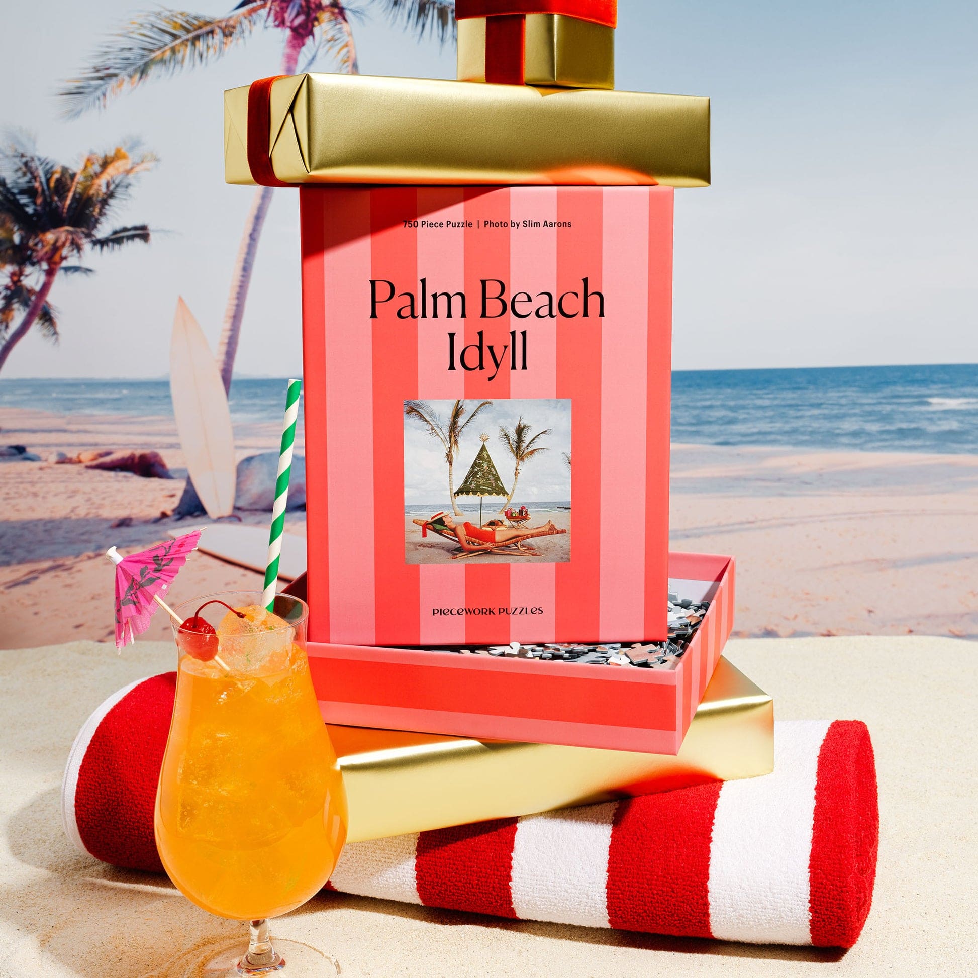 Palm Beach Idyll 750-piece jigsaw puzzle box in coral and pink stripes with tropical beach scene, styled with cocktail and towel