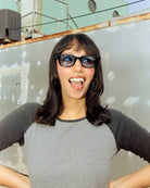 Woman wearing sunglasses and a gray shirt on a rooftop with a metal wall and sky in the background.