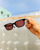 Sunglasses with red lenses held by a hand against a blurred outdoor background