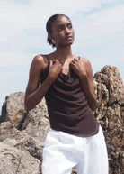 Woman wearing dark brown satin camisole with thin straps and white pants, standing outdoors against rocky backdrop