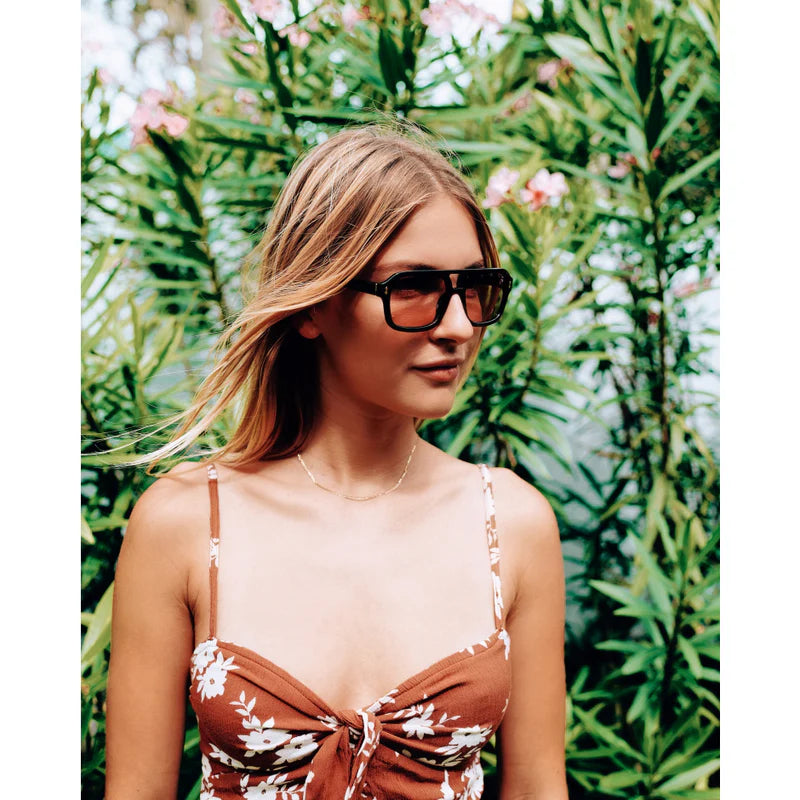 Woman wearing sunglasses and a floral dress in front of green foliage