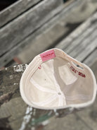 White baseball cap with pink thriftkind label tag visible on interior lining, displayed upside down on wooden surface