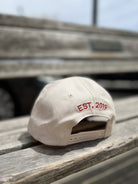 White baseball cap with red EST. 2019 embroidery on back, adjustable strap closure, displayed on wooden bench outdoors