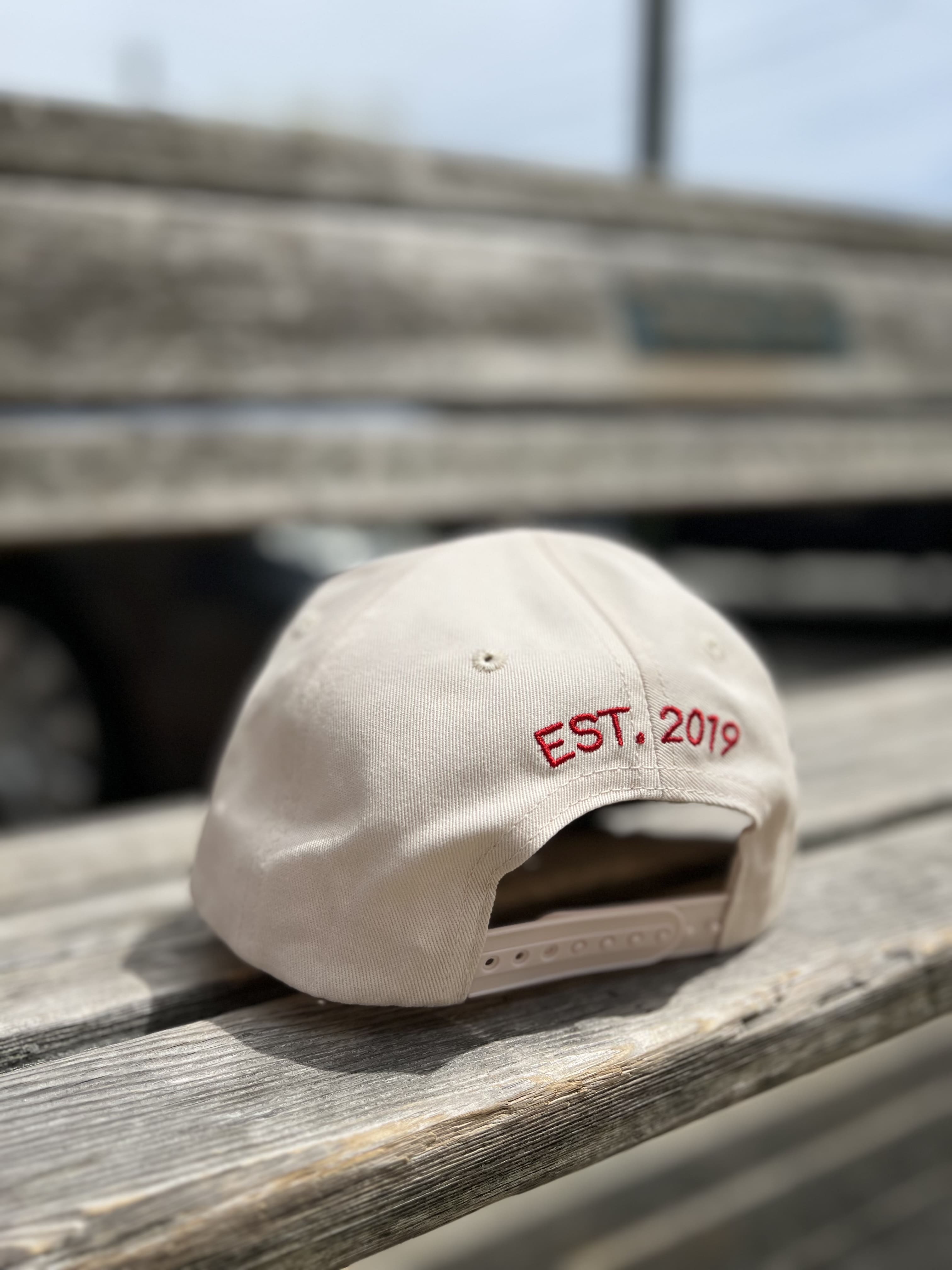 White baseball cap with red EST. 2019 embroidery on back, adjustable strap closure, displayed on wooden bench outdoors