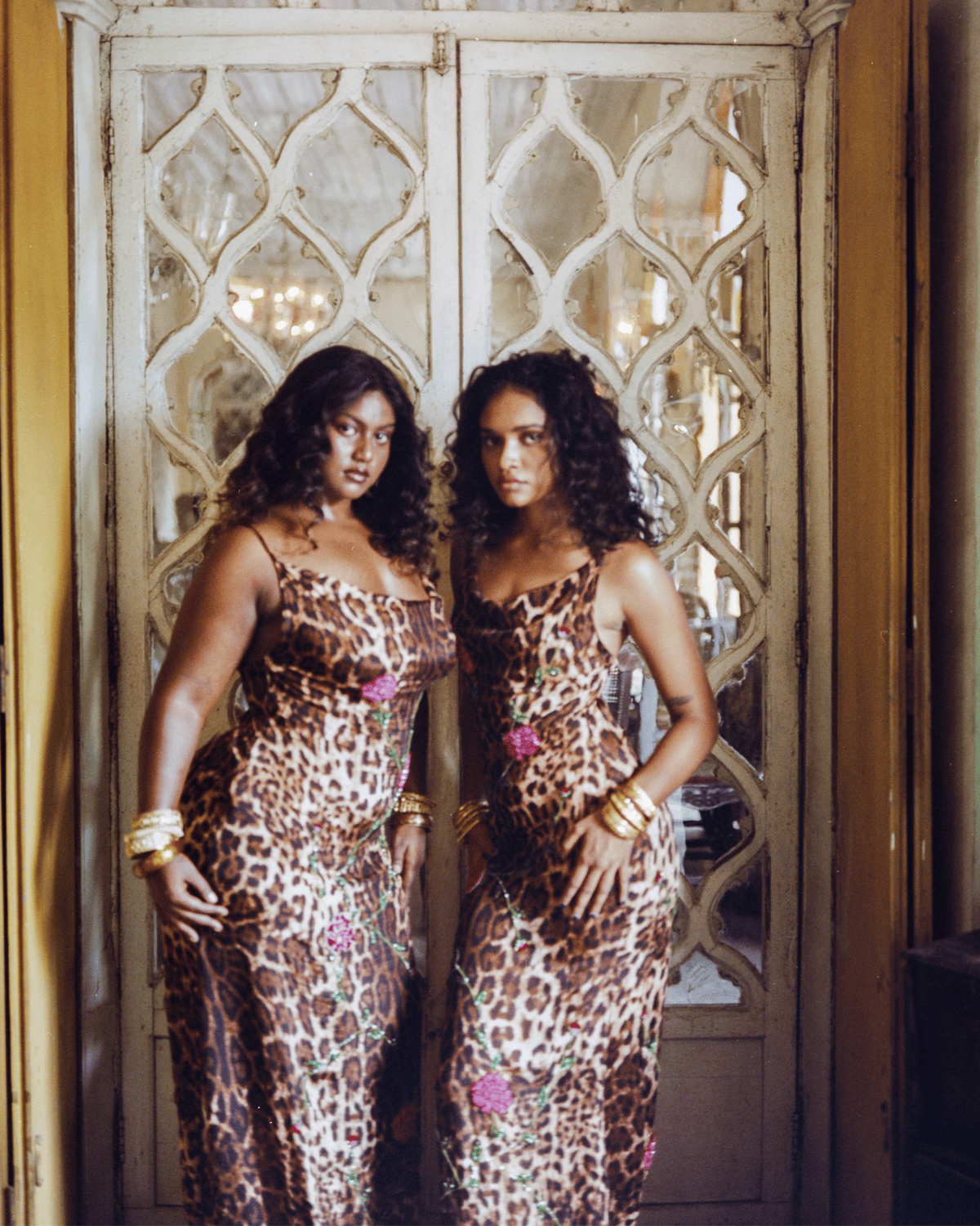 Two women in leopard print slip dresses with floral embroidery and gold cuff bracelets standing by ornate mirrored doors