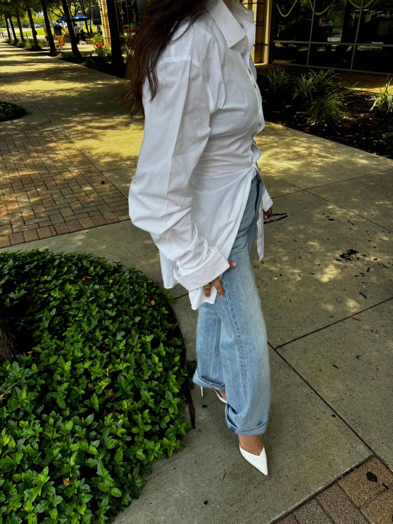 Woman wearing oversized white button-down shirt with light wash denim jeans and white pointed-toe heels in outdoor setting
