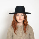 Black wide-brim fedora hat worn by woman with red hair and beige sweater against white background