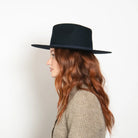 Black wide-brim felt fedora hat with flat crown, modeled on woman with long auburn hair wearing beige sweater, side view