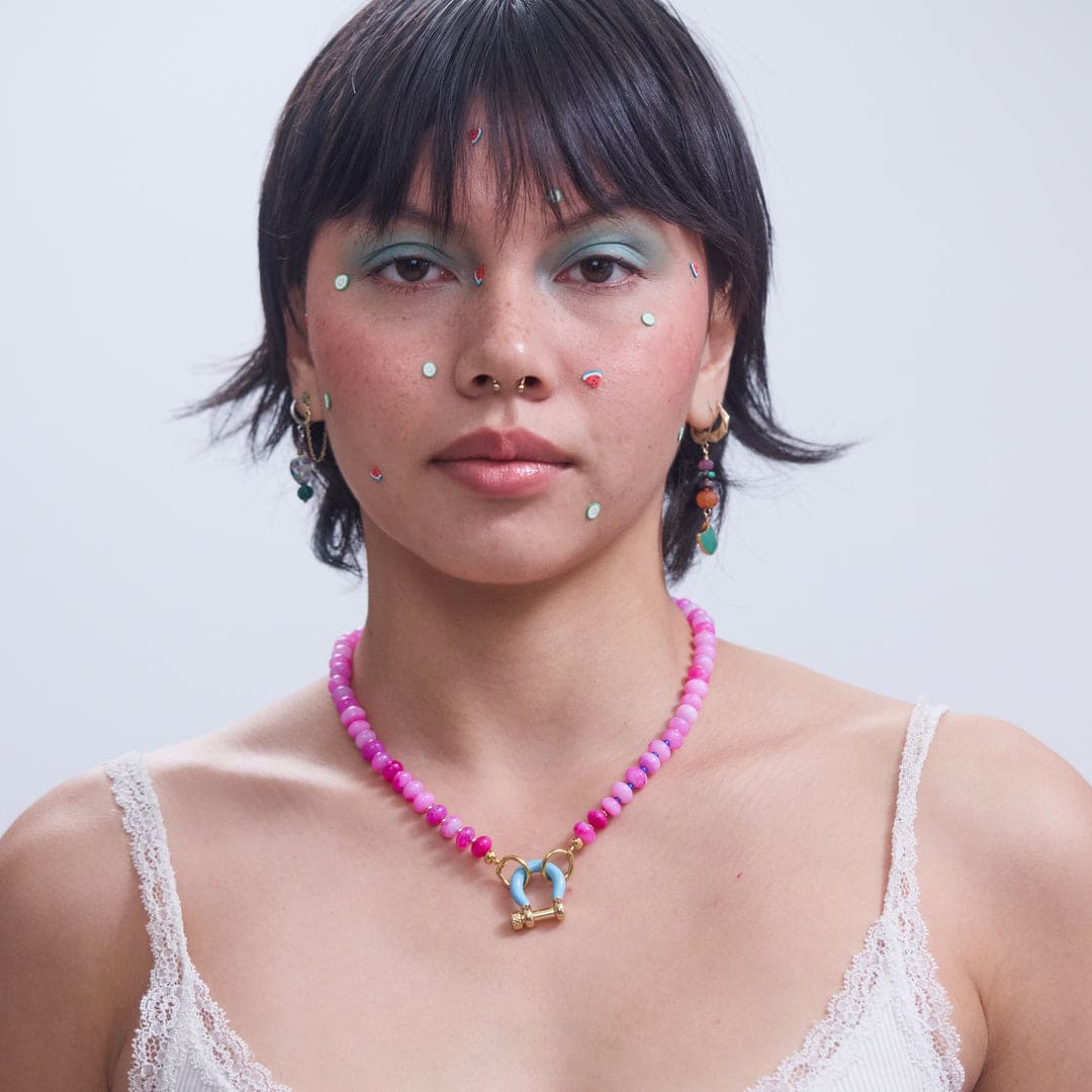Pink beaded necklace with blue enamel horseshoe charm and gold toggle clasp, worn by model with colorful earrings