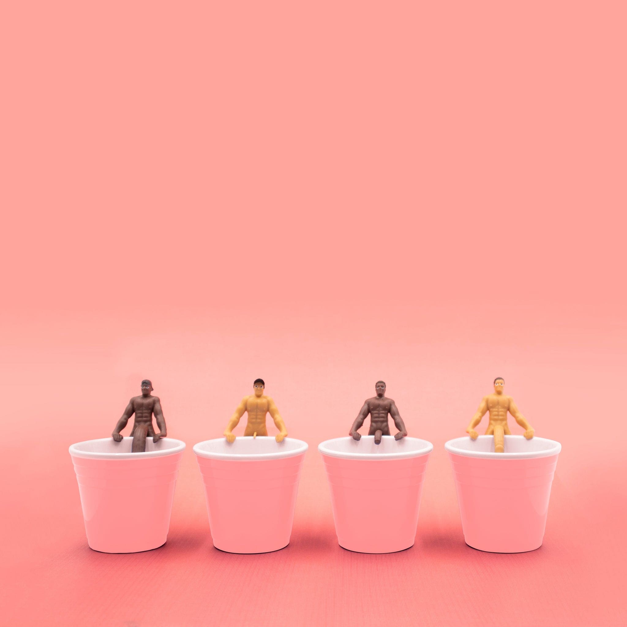 Four shirtless male figurines in brown and yellow tones seated in pink cups against a pink background