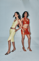 Two models in sequined sets - one in yellow crop top with midi skirt, one in red two-piece with fringe detail, against white backdrop