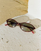 Burgundy red oval sunglasses with gradient lenses on textured white concrete surface with natural lighting