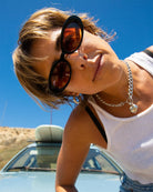 Woman wearing sunglasses and a white tank top in a desert setting with a car in the background.