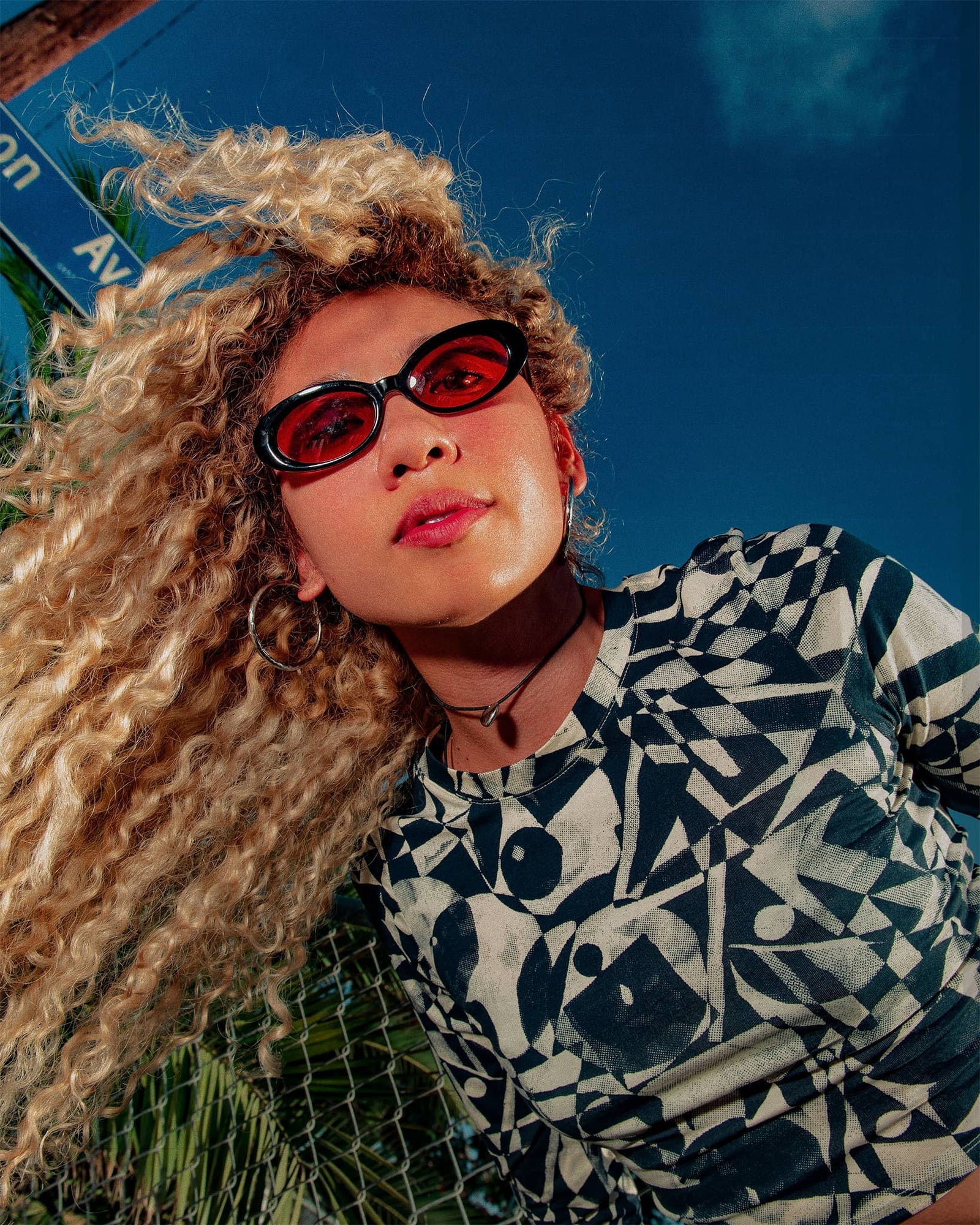 Person with curly hair wearing sunglasses and a patterned shirt against a blue sky.