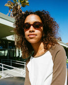 Woman wearing brown oval sunglasses with tortoiseshell frames outdoors in natural sunlight with palm trees in background