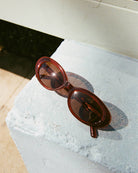 Translucent brown oval sunglasses resting on a textured white concrete surface with dramatic shadow cast by natural light