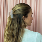 White floral hair claw clip with pearl embellishments holding blonde hair in a half-up style against pink curtain background