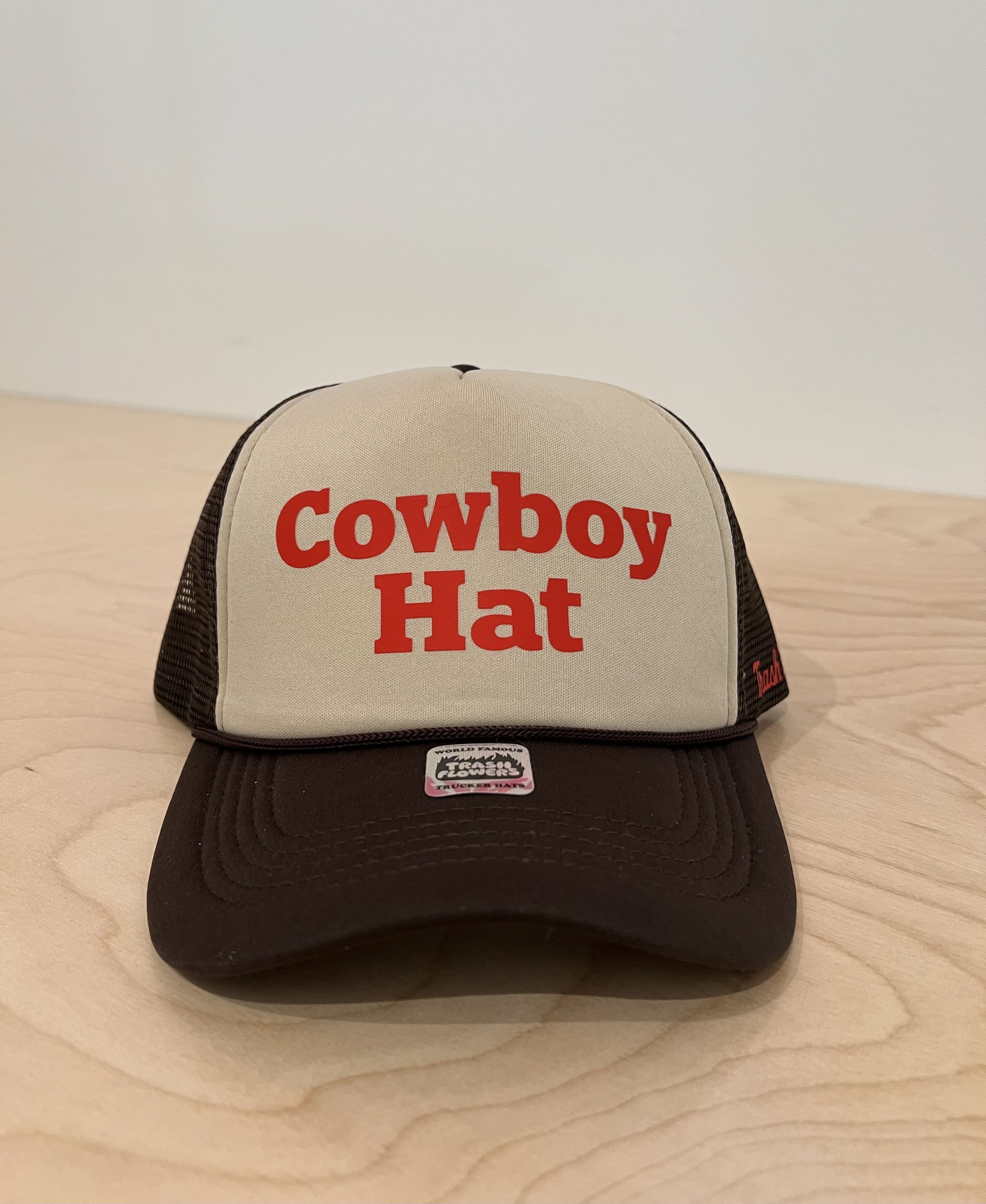 Brown and tan trucker hat with red "Cowboy Hat" text on front panel, mesh back, sitting on wood surface