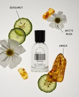 Clear glass perfume bottle with black cap surrounded by bergamot slices, white musk flowers, and amber pieces on beige background